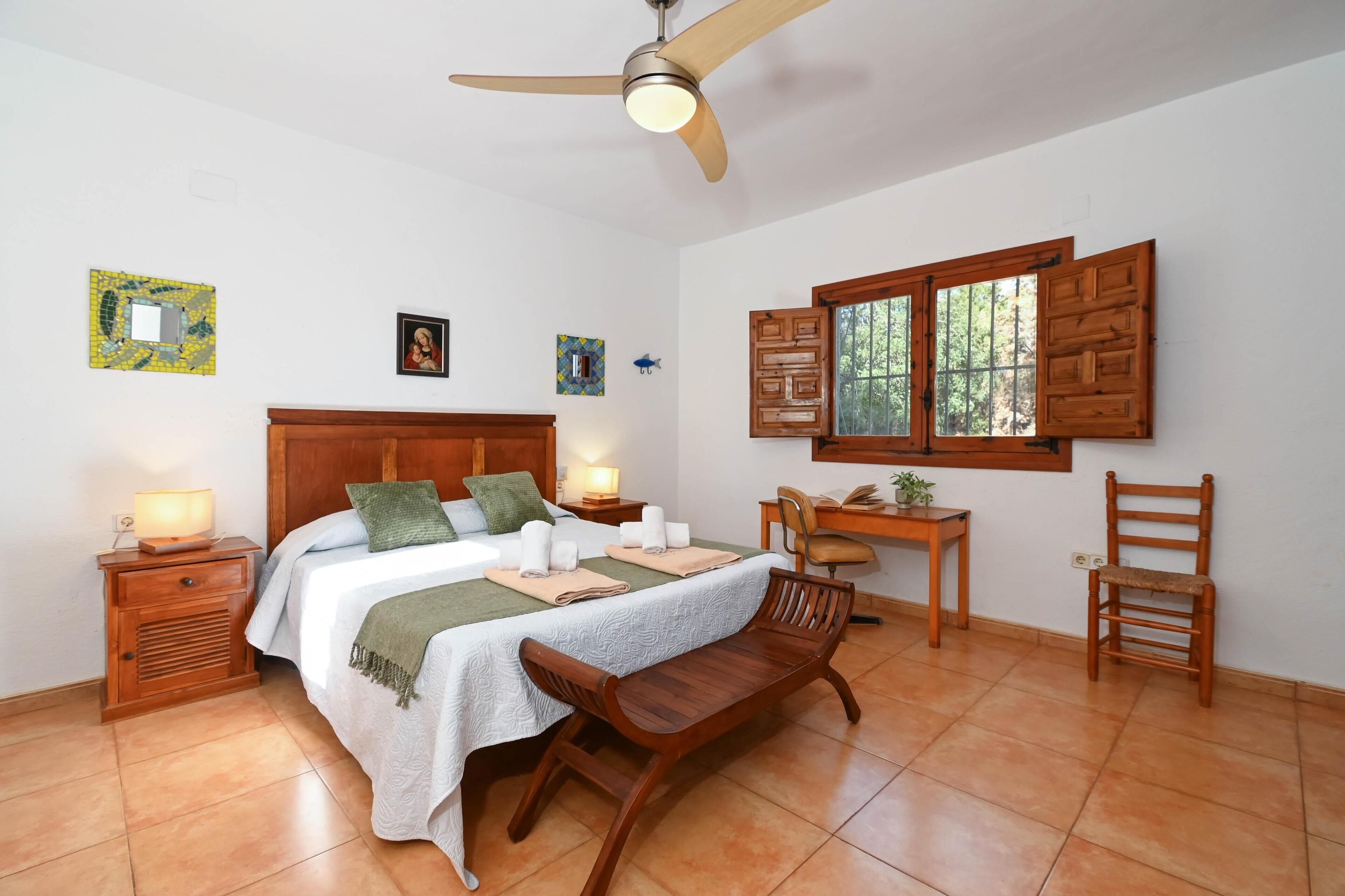 A bedroom with a bed, bedside tables, a desk, a chair, and a window with shutters.