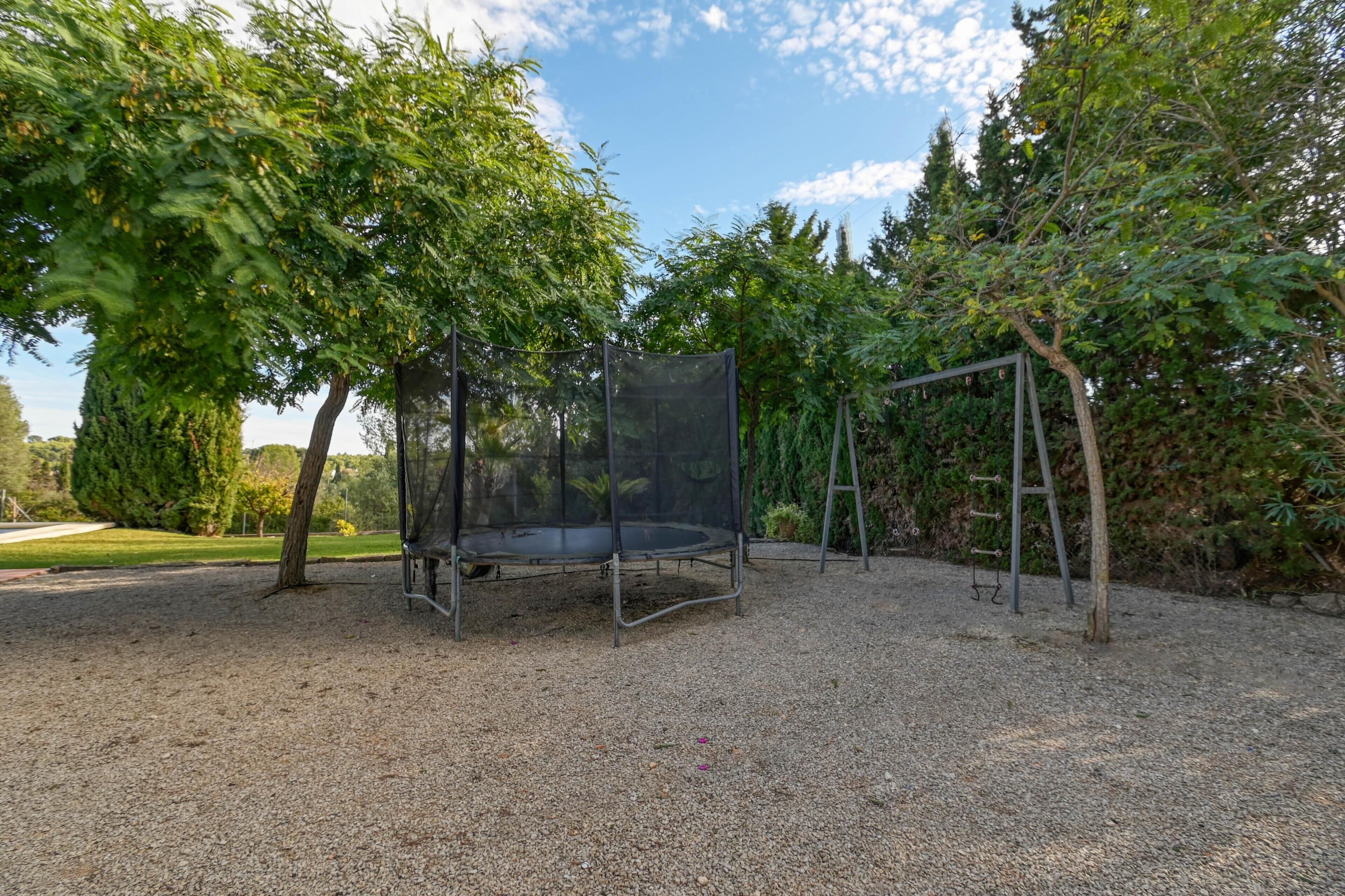 A trampoline with a safety net, surrounded by trees and a gravel area.
