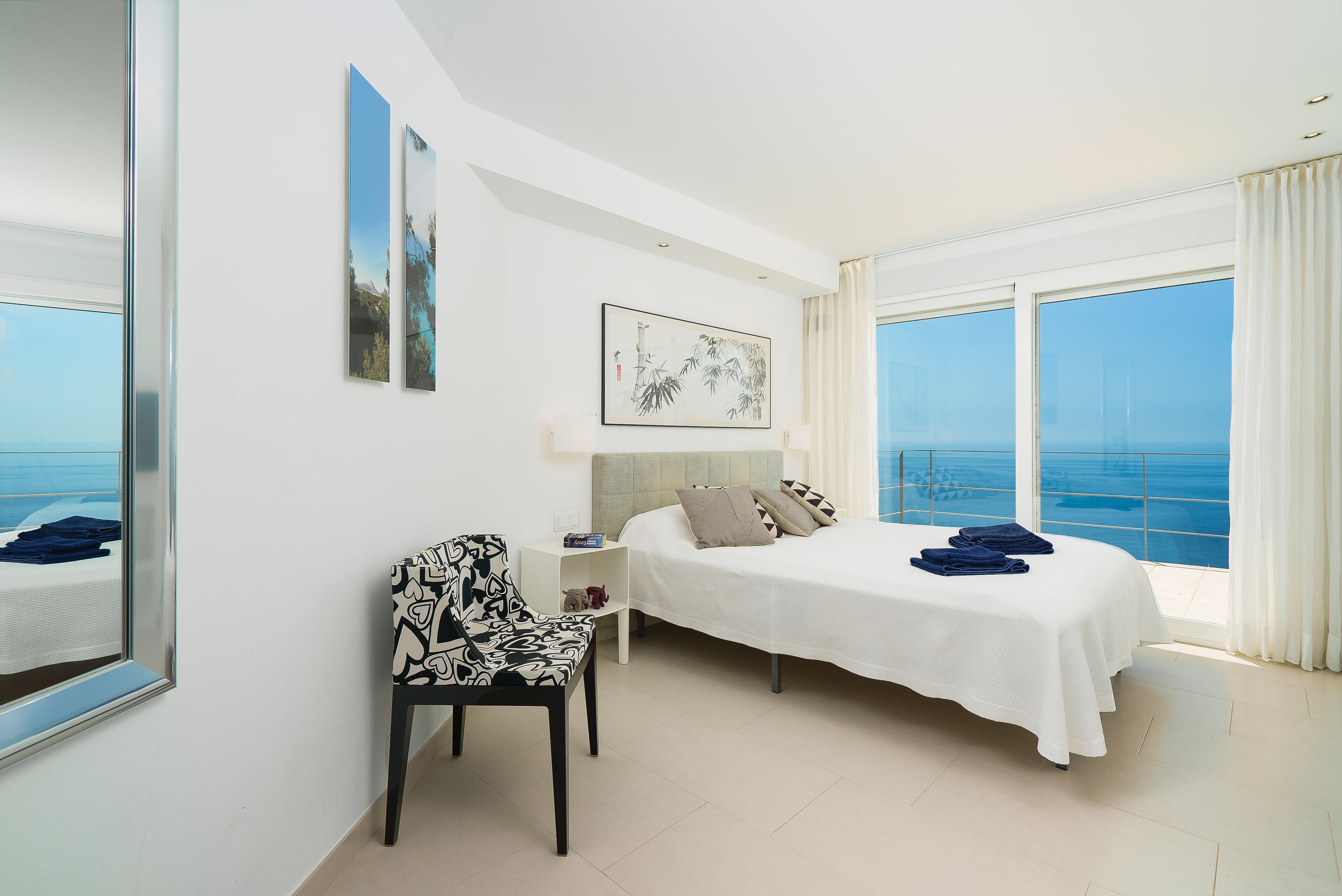 A hotel room with a bed, a chair, a nightstand, and a large window offering a view of the sea.
