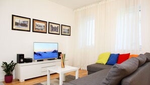 Ein Wohnzimmer mit Flachbildfernseher, einer weißen Unterhaltungsanlage, einem grauen Sofa mit bunten Kissen und drei gerahmten Bildern an der Wand.