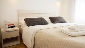 Ein ordentlich bezogenes Bett mit weißer Tagesdecke und zwei dunklen Kissen, ein Nachttisch mit Büchern und einer Lampe sowie ein aufgerolltes weißes Handtuch auf dem Bett.