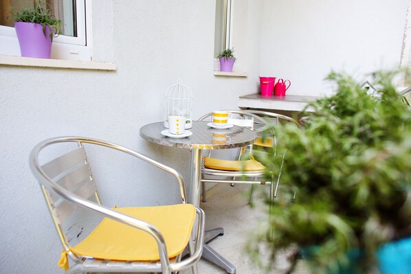 Een klein terras buiten, met een ronde tafel en twee stoelen, een plant in pot en een raam met uitzicht.