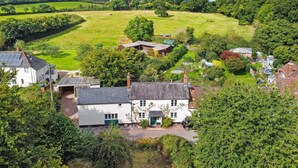 A rural landscape with a white house surrounded by greenery, a clear sky, and rolling hills in the background. - Forge Cottage, Wootton Courtenay (Wootton Courtenay)