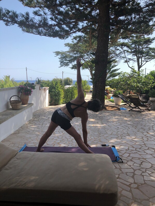 Une personne effectue une posture de yoga à l’extérieur, sur une terrasse, avec vue sur la mer et les arbres.