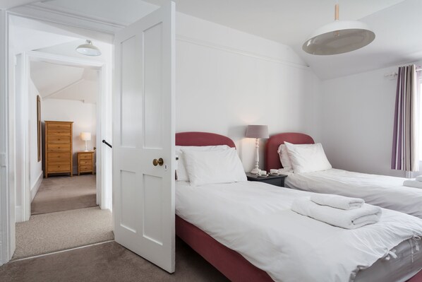 A hotel room with two beds, a wooden dresser, a nightstand with a lamp, and a view of the hallway.
