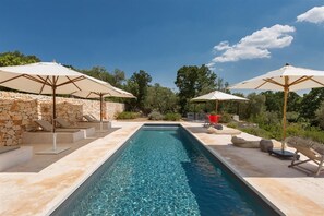 Et rektangulært swimmingpool med klart, blåt vand, omgivet af stenmure og beige gulvfliser. To beige parasoller giver skygge, og der er et par liggestole i syne.