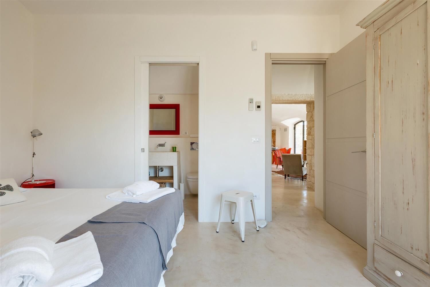 A hotel room with a bed, a desk with a chair, a small stool, a wardrobe, and a bathroom with a mirror and toilet.