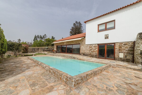 Une piscine en pierre avec une terrasse en pierre, un bâtiment blanc doté d’un toit recouvert de tuiles rouges et un mur en pierre.