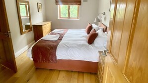 Een slaapkamer met houten vloer, een bed met wit en bruin beddengoed, een nachtkastje, een spiegel, een raam met uitzicht op groen en een hanglamp.