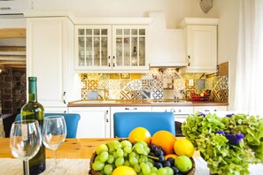 A kitchen with white cabinets, a tiled backsplash, and a wooden table with a fruit bowl and wine glasses.