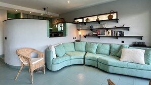 Ein Wohnzimmer mit einem gebogenen, grünen Sofa, einem Korbstuhl und einem Bücherregal mit Büchern und Dekoration.