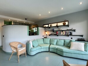 Ein Wohnzimmer mit einem gebogenen, grünen Sofa, einem Korbstuhl und einem Bücherregal mit Büchern und Dekoration.