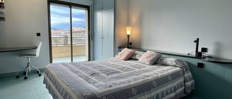 A bedroom with a bed, a desk, a chair, a nightstand, and a window with a view.
