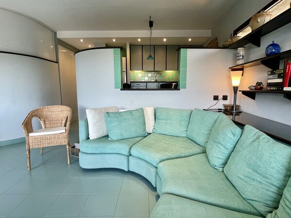 A living room with a curved green sofa, a wicker chair, and a kitchen area with a window above the counter.