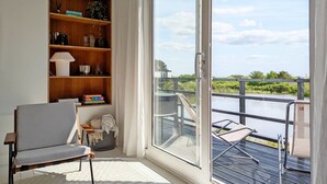 Una stanza con un'ampia porta vetrata scorrevole che dà su un balcone con vista su un corpo d'acqua e vegetazione. Si notano una libreria, una sedia e un tavolino con un tappeto.