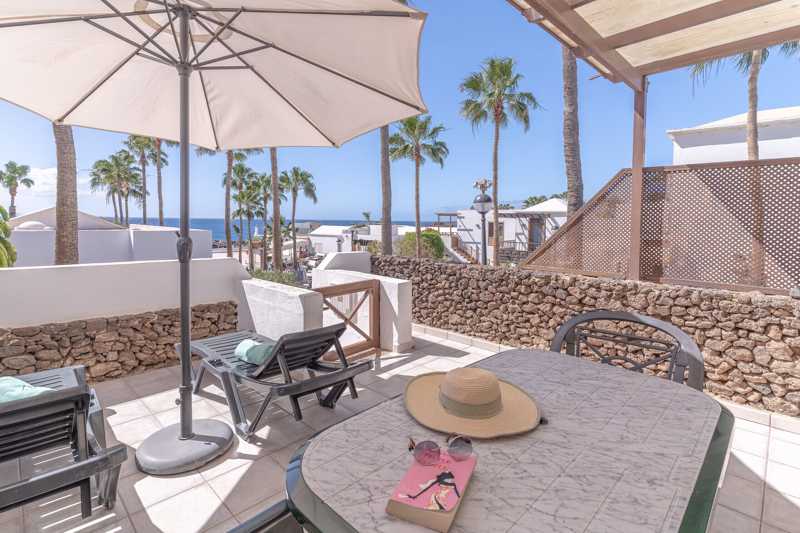 A terrace with a table, chairs, a large umbrella, and a view of the sea and palm trees.
