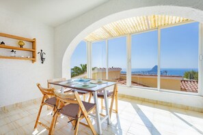 Ein sonnendurchflutetes Zimmer mit Tisch und Stühlen, großen Fenstern mit Blick auf das Meer und ein Berg, sowie ein Regal mit Dekoration.