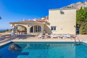 A poolside area with a clear pool, sun loungers, a dining area, and a building with a tiled roof. - Villa Alicia - Plus (Calpe)