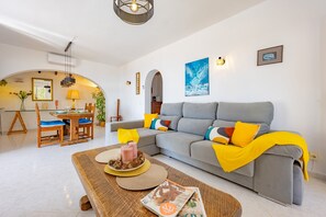 Ein modernes Wohnzimmer mit grauem Sofa, gelben Kissen, einem Holztisch mit Zeitschriften und einem Essbereich mit Holztisch und Stühlen.