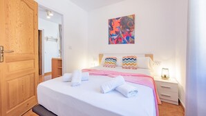 Hotellihuone, jossa on puinen ovi, valkoisilla ja vaaleanpunaisilla tekstiileillä varustettu sänky, värikäs abstrakti maalaus seinällä ja yöpöytä, jossa on lamppu.