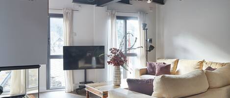 A modern living room with a flat-screen TV, a sofa with cushions, a wooden coffee table, and a vase with plants.