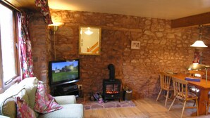A cozy living room with a stone wall, a television, a fireplace, and a dining area with wooden furniture. - Allerford Cottage, Near Dunster (Near Dunster)