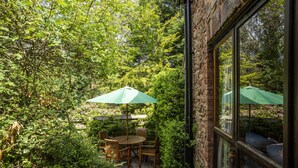 A patio with a round table and chairs, surrounded by lush greenery and a brick wall. - Allerford Cottage, Near Dunster (Near Dunster)