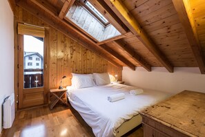 A bedroom with a wooden ceiling, a skylight, a bed with white linens, a bedside table with a lamp, and a view of a building outside.