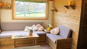 A cozy cabin interior with a wooden bench and a sofa, a small table with a bottle and glasses, and a window with a view of greenery. - 2 bedroom luxury lodge with heated covered pool (Henansal)