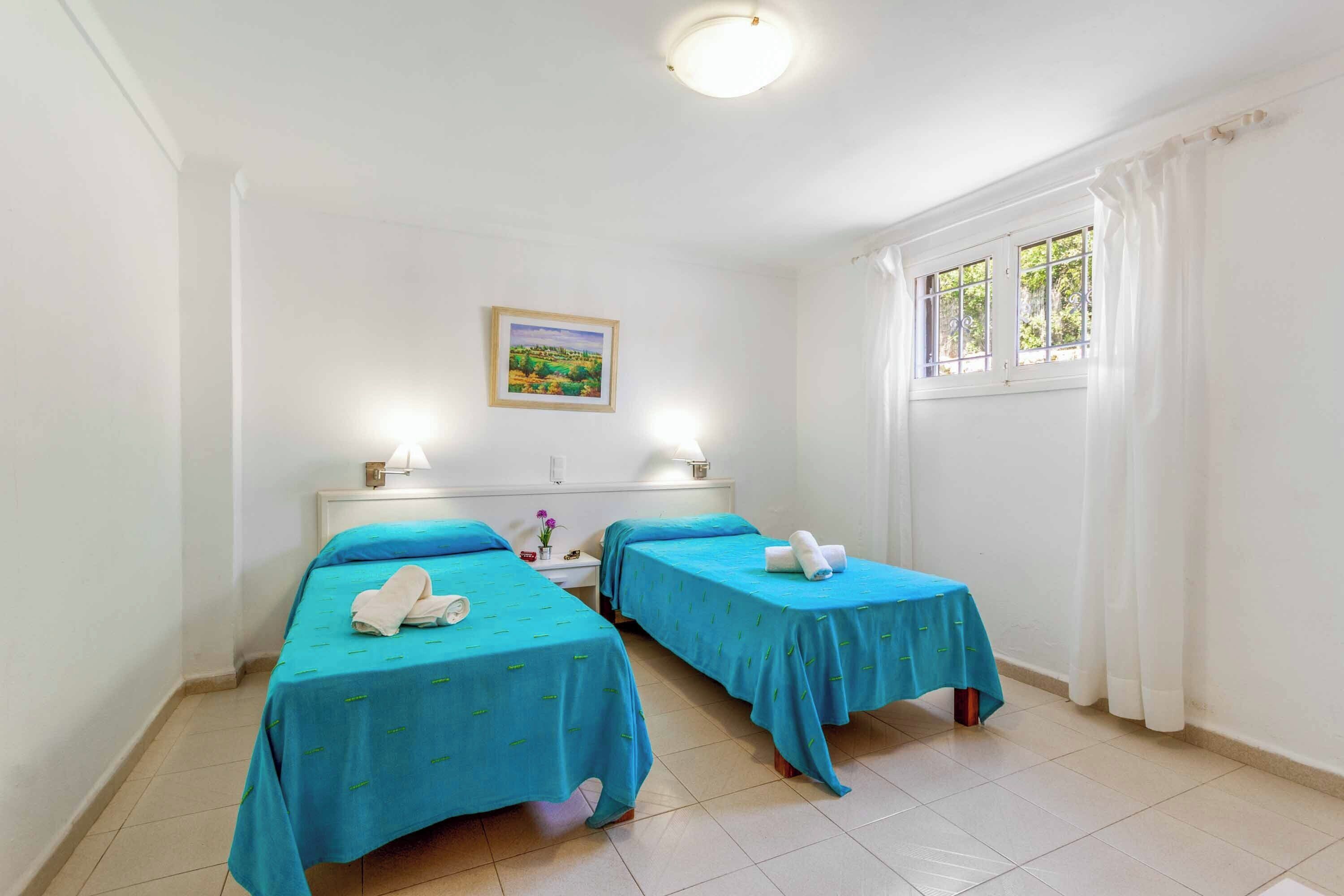 Une chambre avec deux lits, chacun recouvert d’une couverture bleue et orné d’une serviette blanche sur l’un des lits. On y trouve une table de chevet avec une lampe et un tableau encadré accroché au mur.