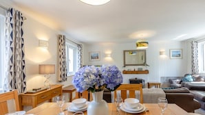 Un comedor con una mesa de madera dispuesta para cuatro, un jarrón de flores y un sofá al fondo.