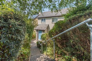 A pathway leading to a house with a blue door, surrounded by dense foliage and a metal handrail. - 3 Bed in Braunton (oc-salar) (Georgeham)