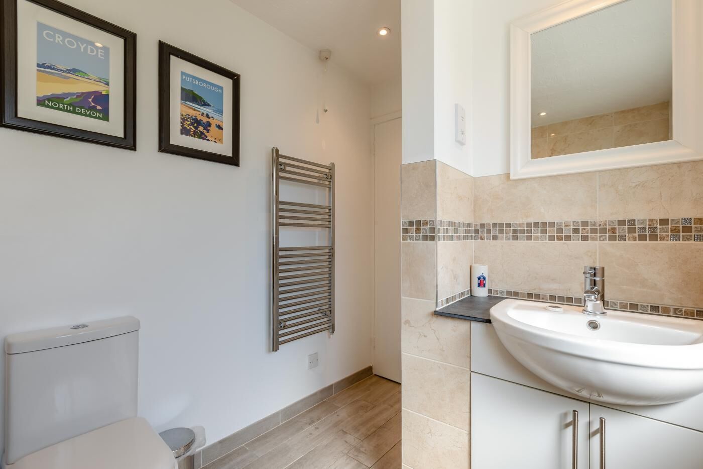 A bathroom with a toilet, a towel rack, a sink, and a mirror. There are two framed pictures on the wall.