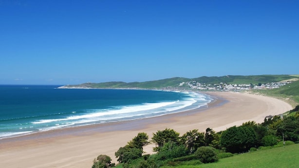 Un paisaje costero con playa arenosa, olas oceánicas y un pueblo al fondo.