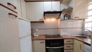 Cocina compacta con armarios blancos, horno integrado, lavavajillas y una encimera con un hervidor y otros objetos pequeños.