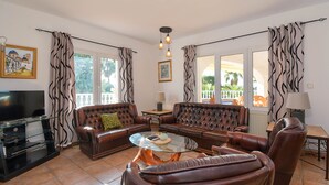 Ein Wohnzimmer mit Ledersofas, einem Glastisch, einem Flachbildfernseher und großen Fenstern mit Vorhängen.