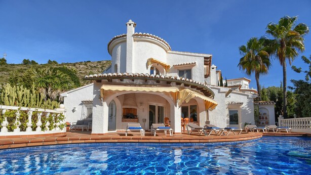 Blick vom Pool auf eine Villa im mediterranen Stil, mit Pool, Sitzgelegenheiten im Freien und Palmen.