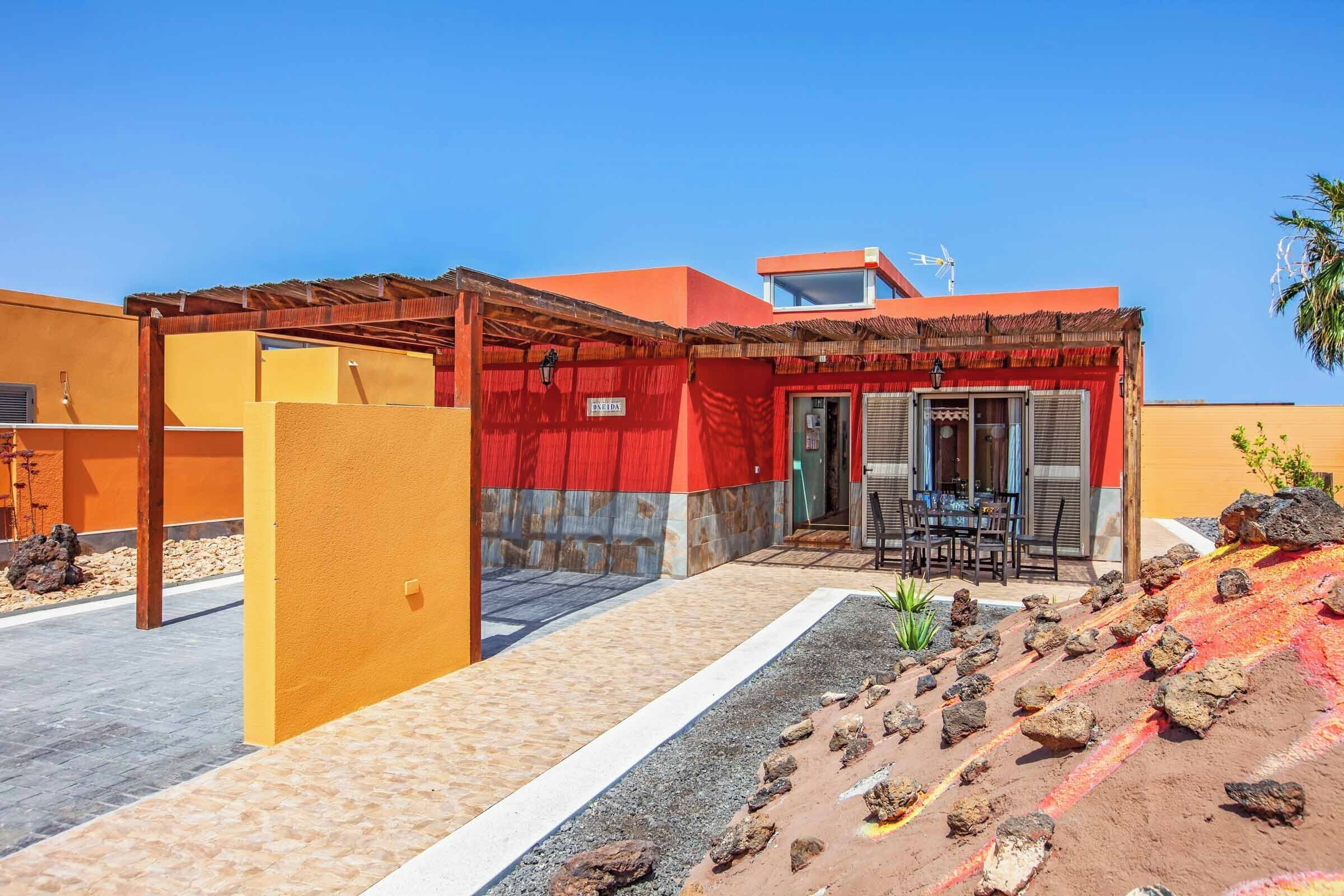 Un bâtiment moderne à la façade rouge et jaune, doté d’un patio couvert et d’un petit espace extérieur aménagé pour s’asseoir.