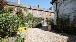 A stone building with a gravel path leading to a gated entrance, surrounded by greenery and potted plants. - Grace Cottage, Porlock (Porlock)
