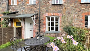 A stone house with a small patio, a table with chairs, and a bottle of wine. - Grace Cottage, Porlock (Porlock)