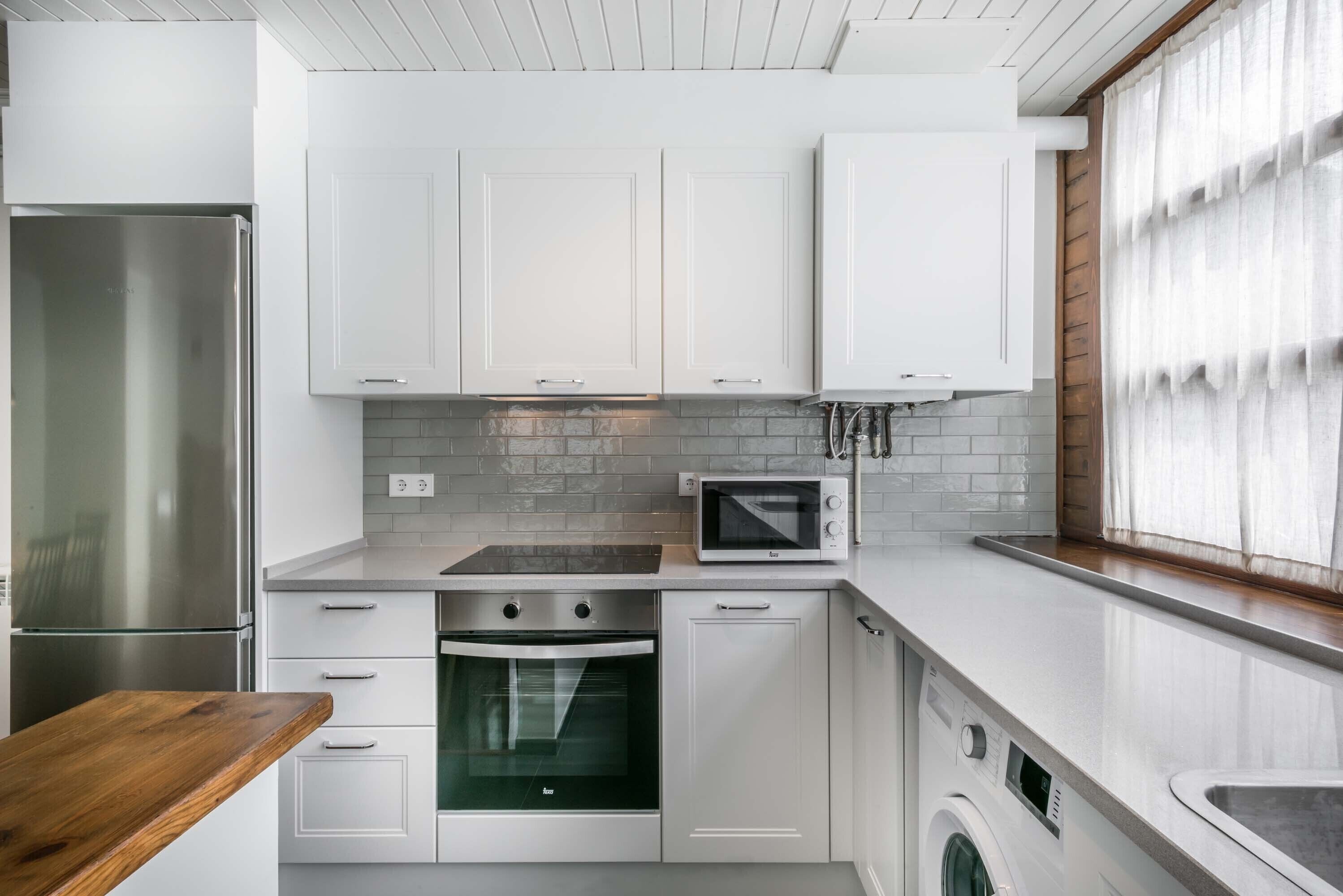 Eine moderne Küche mit weißen Schränken, einem Edelstahlkühlschrank, einer Mikrowelle, einem Backofen und einer Waschmaschine.