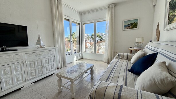 A modern living room with a flat-screen TV, a white cabinet with decorative panels, a striped sofa, and a view of the outdoors through large windows. - APARTAMENT GRECIA (Estartit)