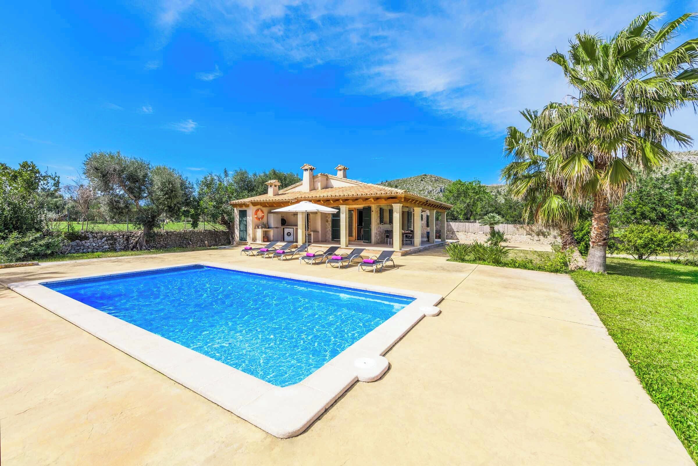 Blick auf einen Pool vor einer Villa im mediterranen Stil, mit Liegestühlen und einer Palme.