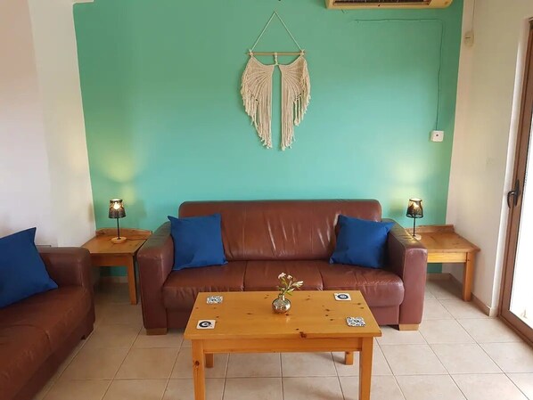 A living room with a brown leather sofa, two blue cushions, a wooden coffee table with a vase, and a macrame wall hanging.