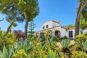 A two-story house with a white facade and a tiled roof, surrounded by various plants and trees.