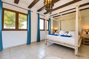 A four-poster bed with white linens and blue accents, a wooden ceiling with exposed beams, and a view of trees through the windows.