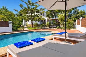 A swimming pool with lounge chairs and an umbrella, surrounded by greenery and a building with a white facade.