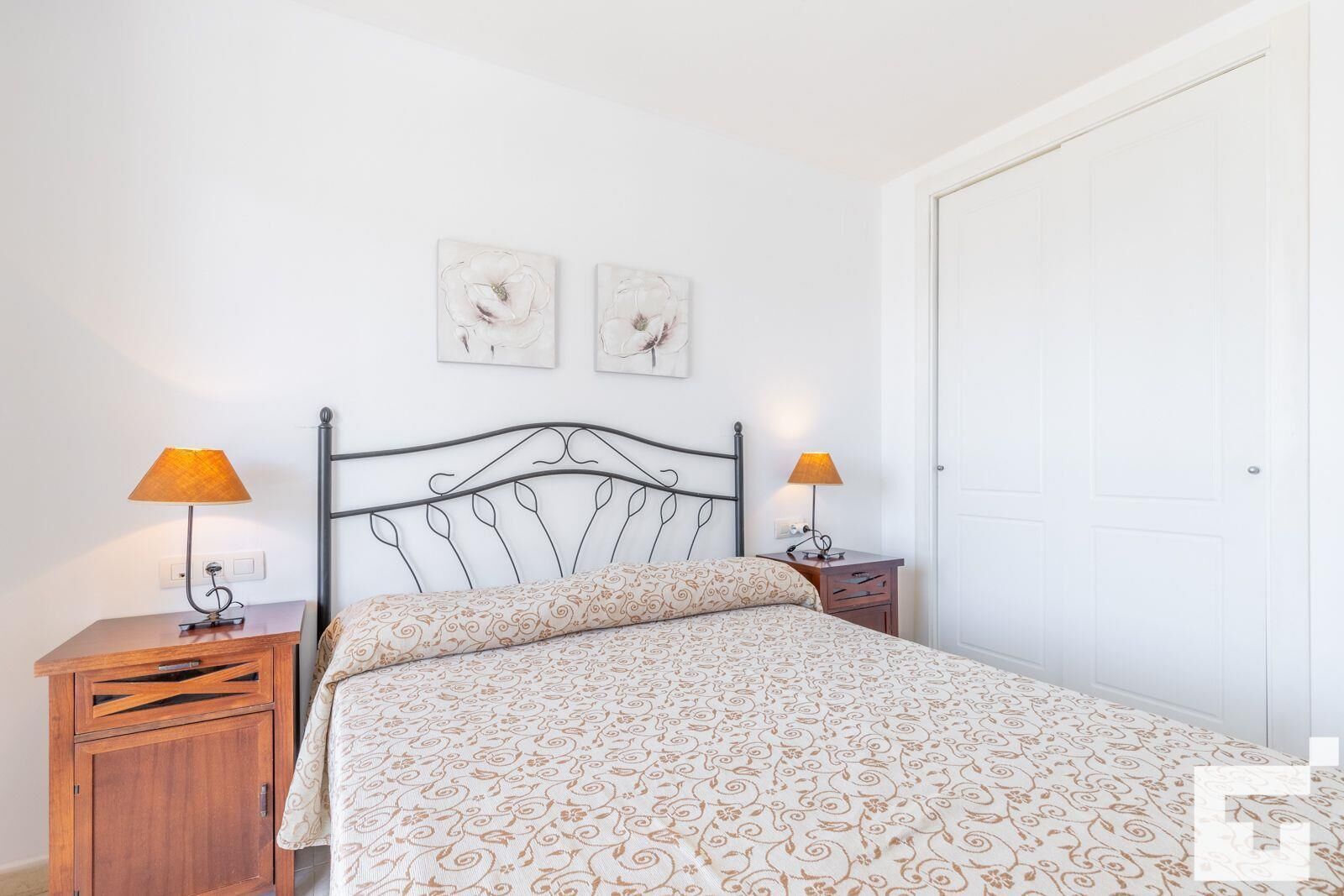 Un dormitorio con estructura de cama metálica, dos mesitas de noche con lámparas, un armario de madera y dos cuadros florales colgados en la pared.