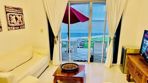 A living room with a white sofa, a wooden coffee table, and a television on a wooden cabinet. A sliding glass door leads to an outdoor patio with a view of the sea.