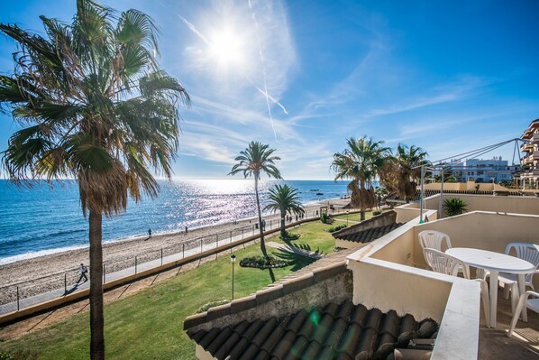 A balcony with a view of the beach and ocean, palm trees, and a white table and chairs.